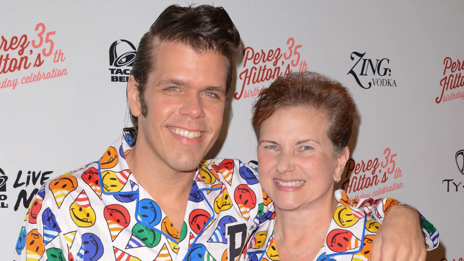 Perez Hilton and an older woman smile, both wearing white shirts patterned with colorful smiley faces in party hats, at an event.
