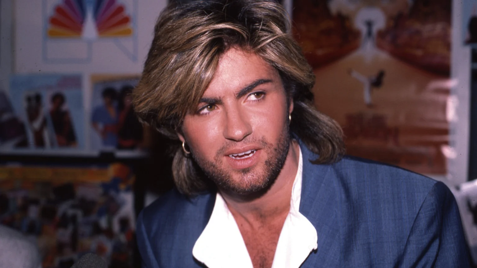 George Michael, with brown hair, beard, and an earring, wears a blue suit jacket and white shirt, looking slightly right during an event, with blurry posters and an NBC logo in the background.