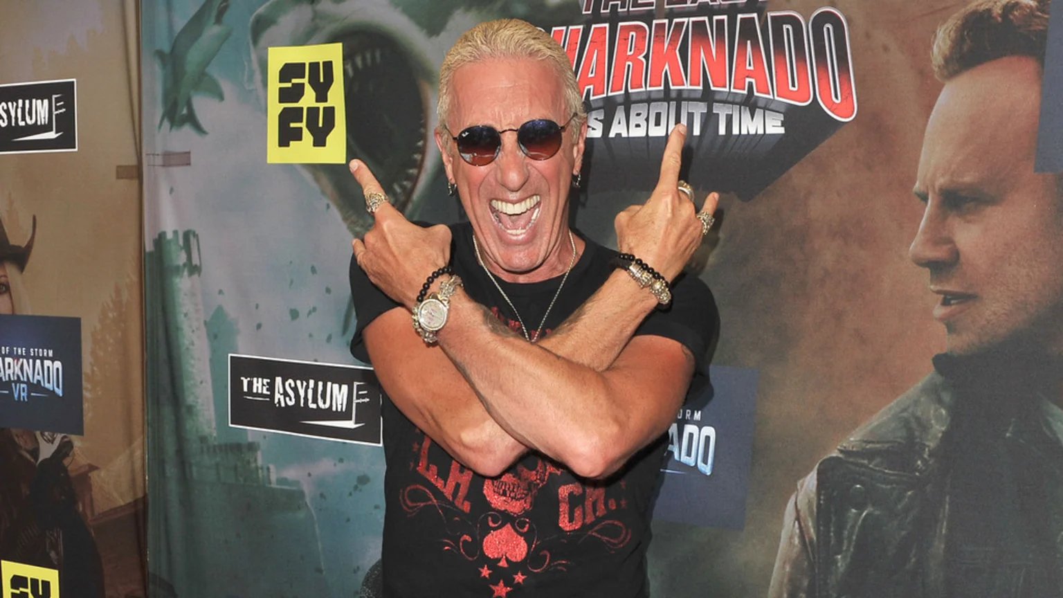 Close-up of musician Dee Snider making 'rock on' gestures, wearing sunglasses and jewelry, standing before banners promoting 'The Last Sharknado: It's About Time' and 'Sharknado VR' with logos for Syfy and The Asylum visible.