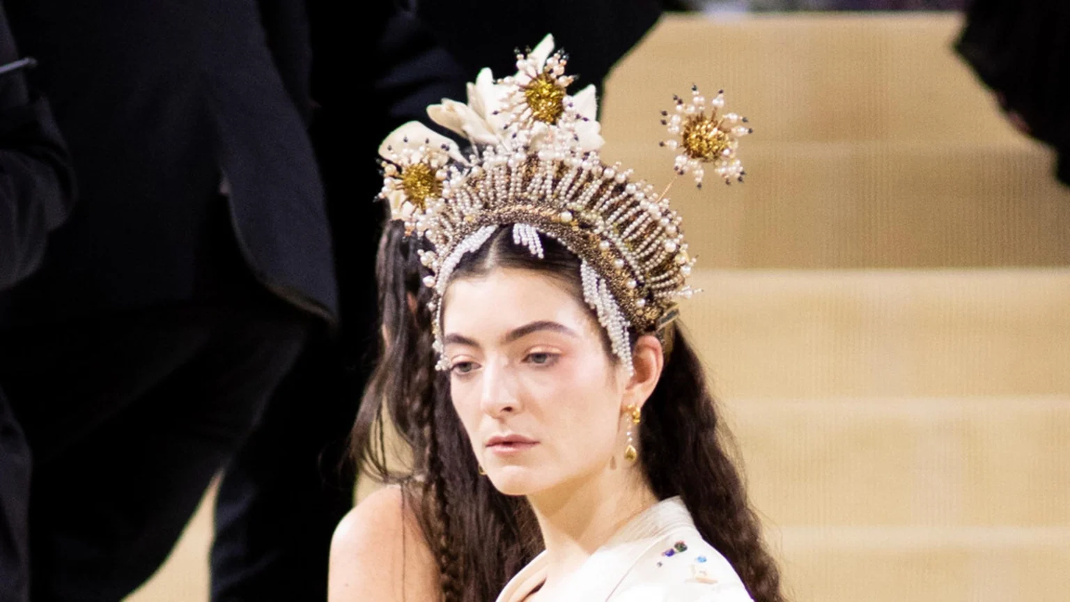 Lorde is seen in a close-up, wearing an elaborate crown adorned with pearls and golden floral designs, featuring long dark hair and a light-colored outfit, looking down with a contemplative expression.