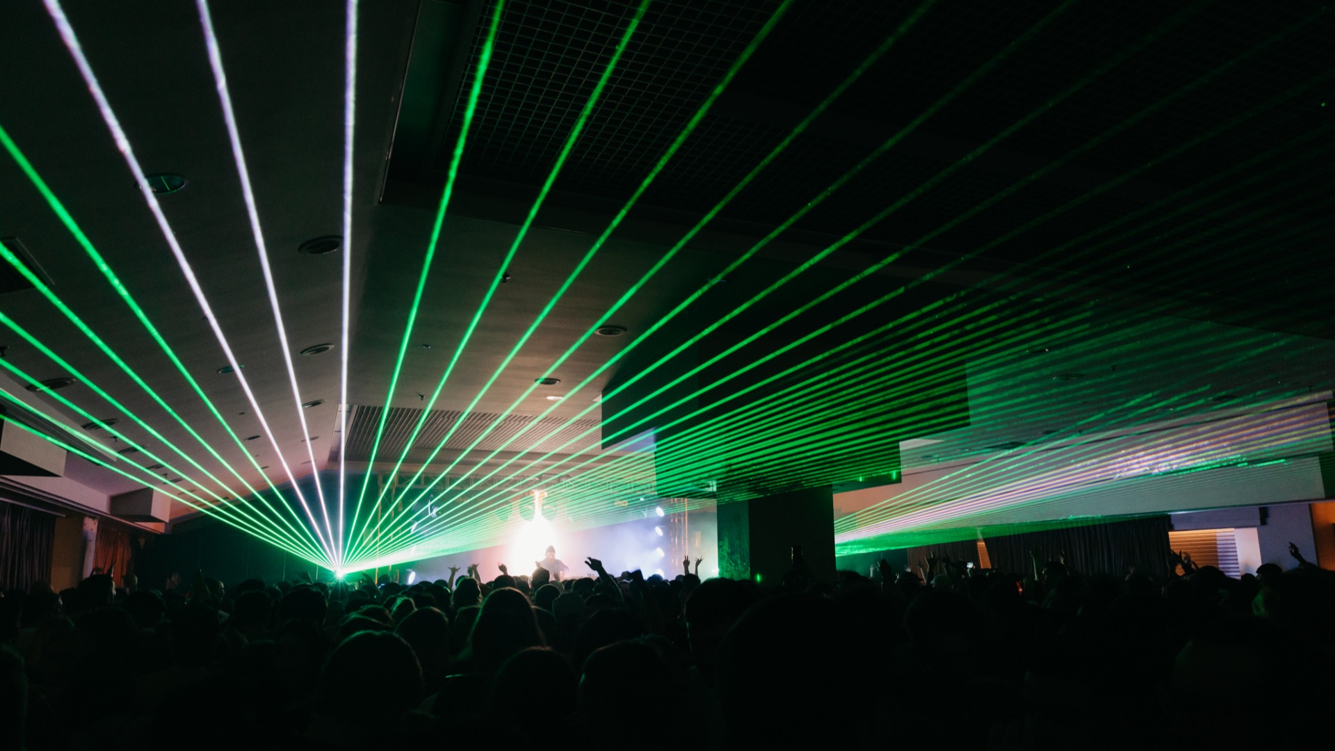 A large crowd of silhouetted people with raised hands at a concert, illuminated by numerous green and white laser beams fanning out from the stage towards the ceiling.