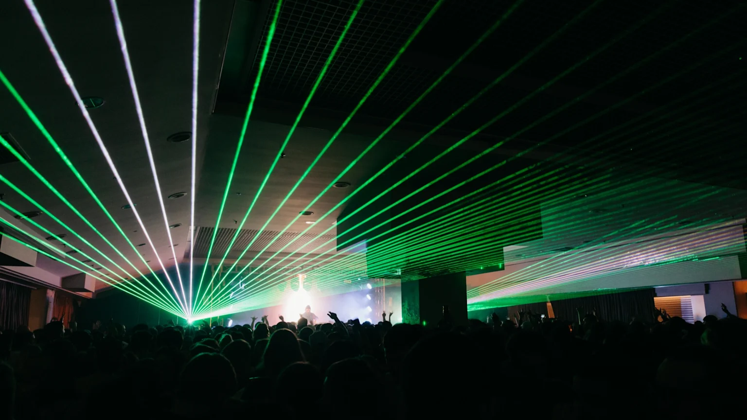 A large crowd of silhouetted people with raised hands at a concert, illuminated by numerous green and white laser beams fanning out from the stage towards the ceiling.