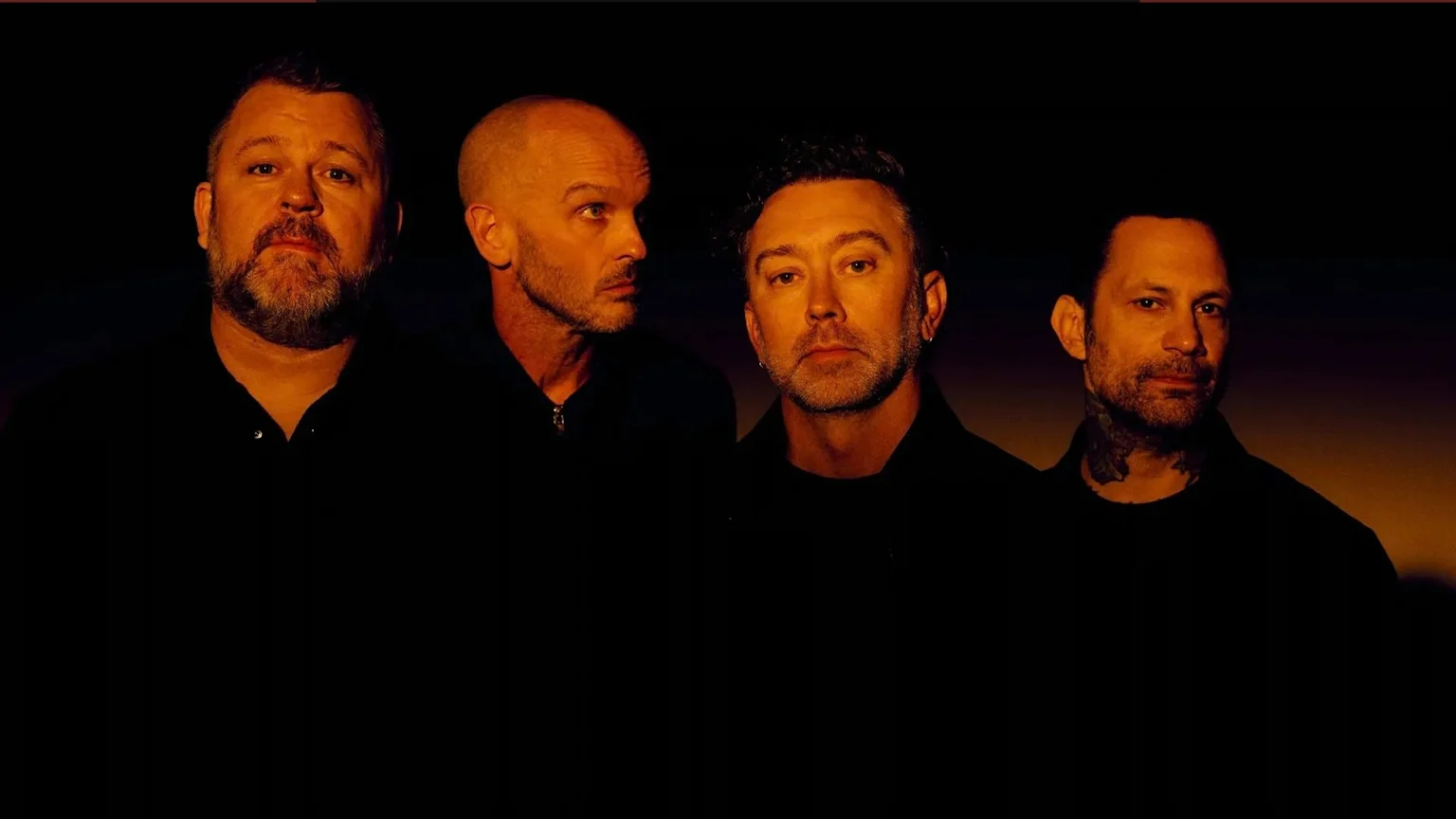 Four men with beards and dark shirts, lit by orange light, are shown from the chest up against a dark background; three face forward, one looks to the left.