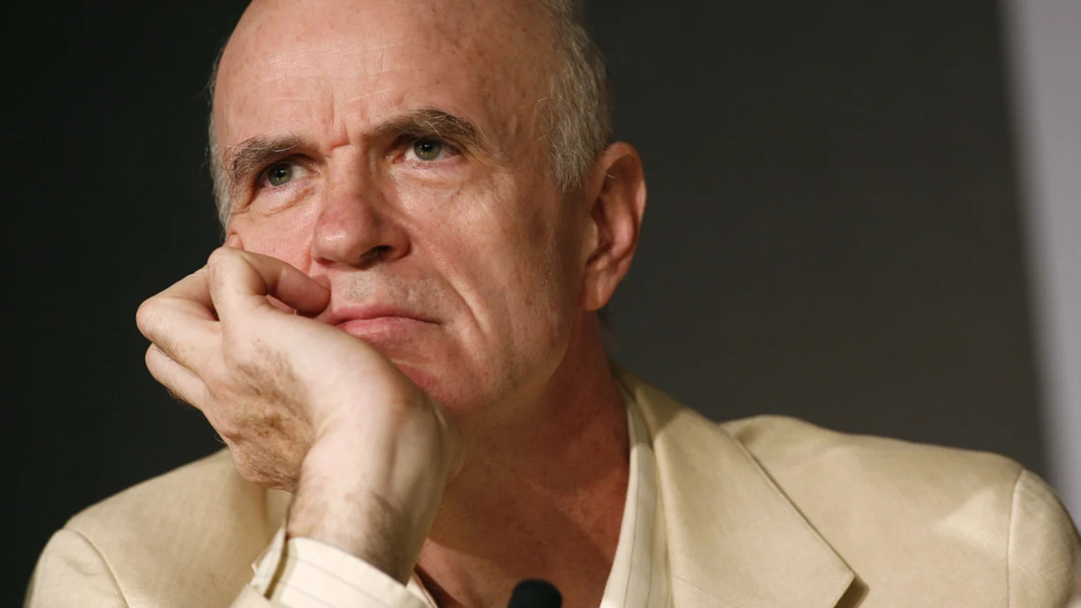 An older man, bald on top with grey hair on the sides, wearing a light-colored jacket, rests his hand on his chin while looking thoughtfully upwards and to the left.