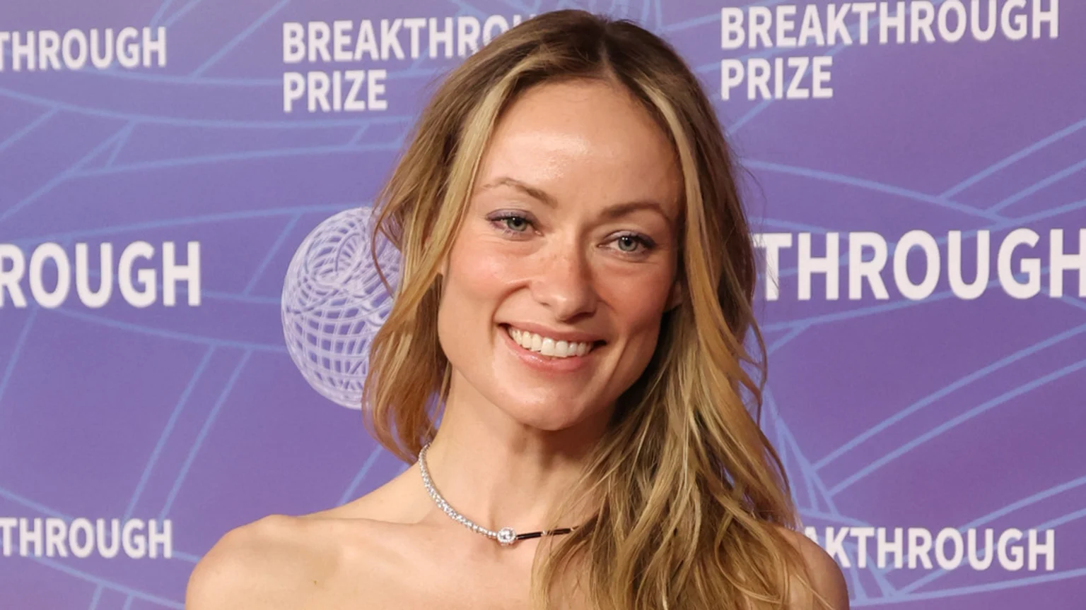 Olivia Wilde smiles at the camera, wearing a strapless top and a diamond necklace against a purple background with "BREAKTHROUGH PRIZE" text.