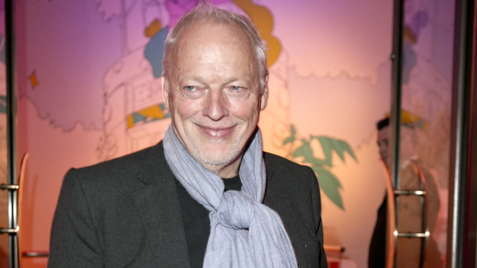 David Gilmour smiles while wearing a dark jacket and light blue scarf, standing in front of a brightly lit wall adorned with colorful cartoon-like illustrations.