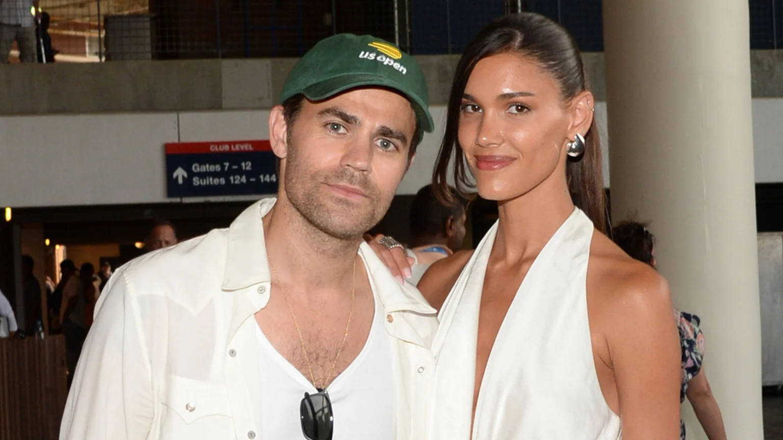 Paul Wesley in a green US Open cap and white shirt, standing next to Natalie Kuckenburg in a white halter top, smiling at the camera at an indoor venue.