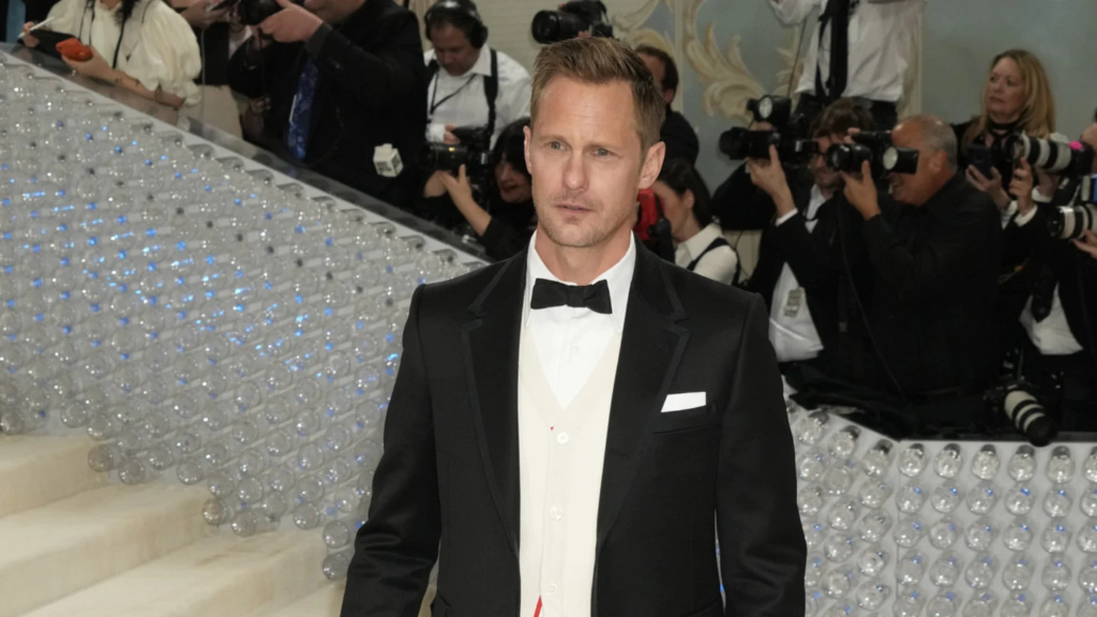 Alexander Skarsgard in a black tuxedo with a white shirt, black bow tie, and light cardigan stands before photographers and a decorative wall of clear spheres with blue lights.