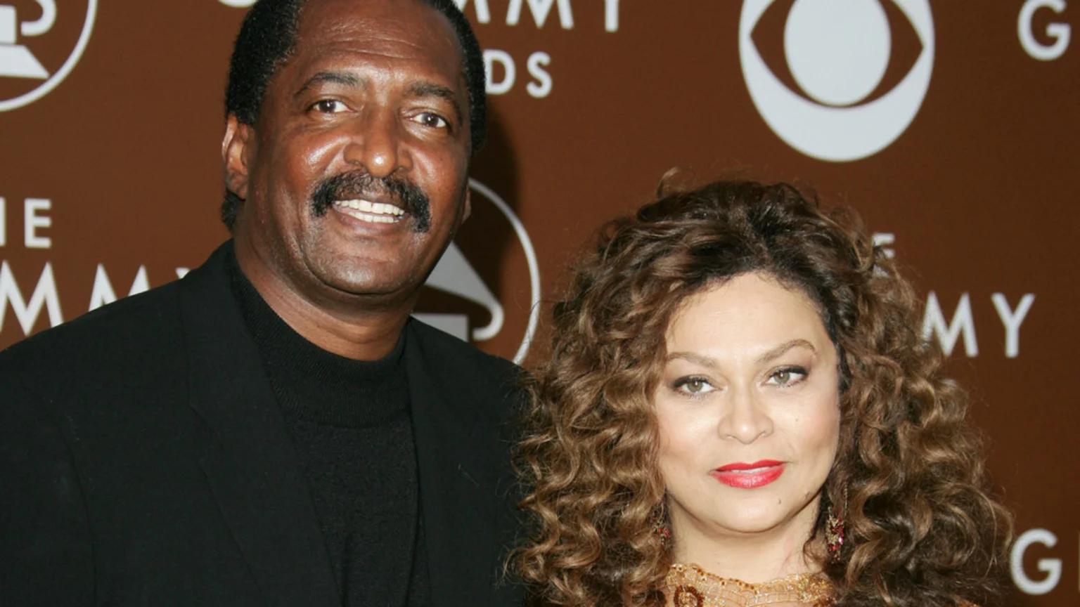 Mathew Knowles and Tina Knowles smile as they pose together; he wears a black jacket and turtleneck, while she has curly brown hair and a gold top, against a brown background with 'GRAMMY AWARDS' text and a CBS logo.