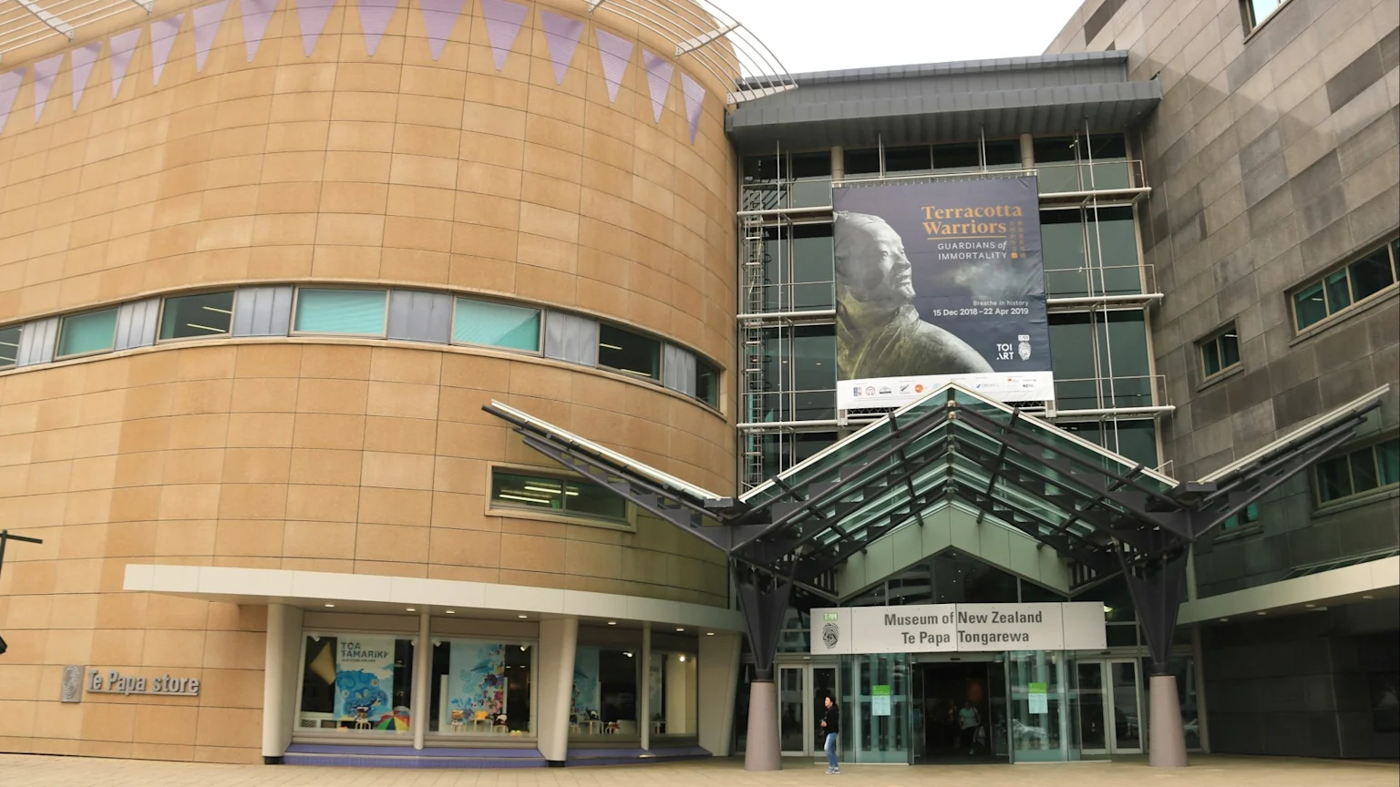 Exterior view of the Museum of New Zealand Te Papa Tongarewa, a large building with a curved tan section and a grey section, featuring a "Terracotta Warriors" banner and a distinctive entrance.