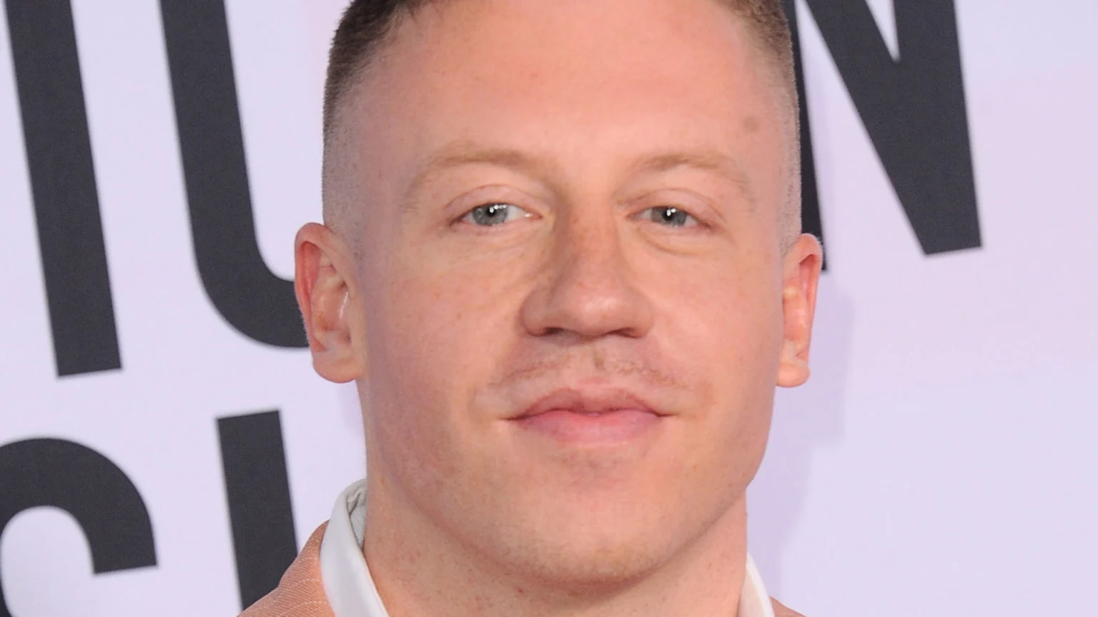 A close-up portrait of a light-skinned man with short, buzzed hair, light blue eyes, and light brown stubble, wearing a light-colored collared shirt and looking directly at the camera with a slight smile.