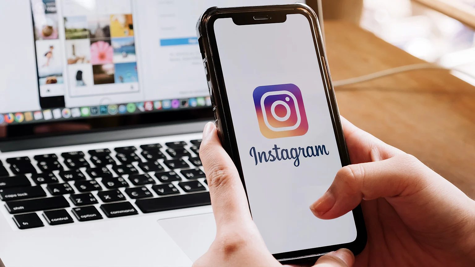 A person holds a black smartphone displaying the Instagram logo and "Instagram" text, with a blurry laptop screen showing an Instagram profile in the background.