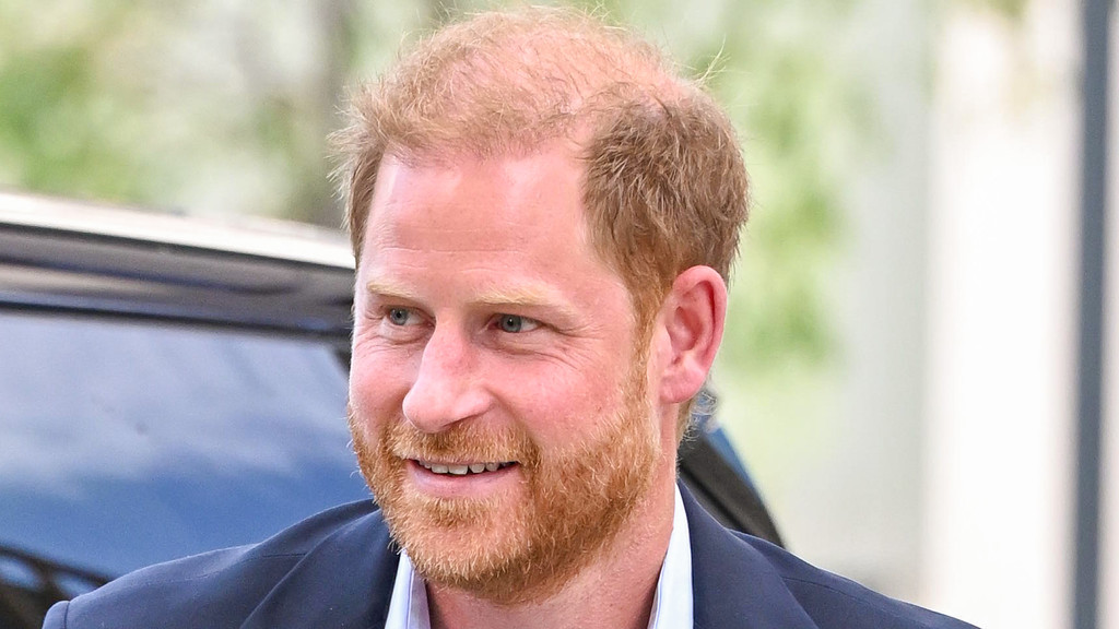 A close-up photo shows Prince Harry smiling, with red hair and a beard, wearing a dark suit jacket and light collared shirt, against a blurred background.