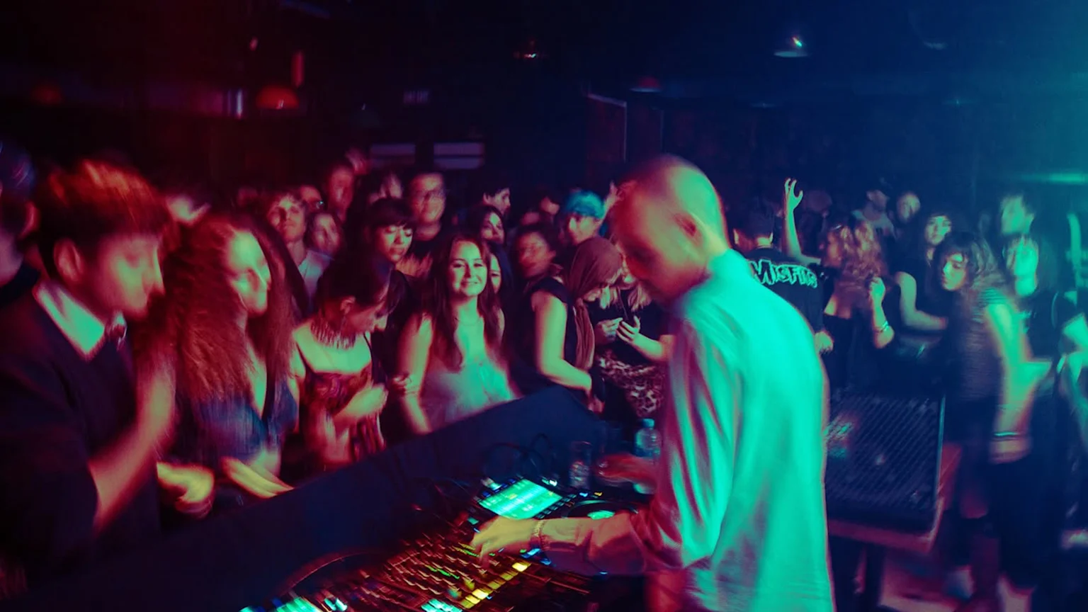 A DJ with a bald head plays music on a mixer in a dark club with a crowd of people under red, blue, and green lights, showing motion blur.