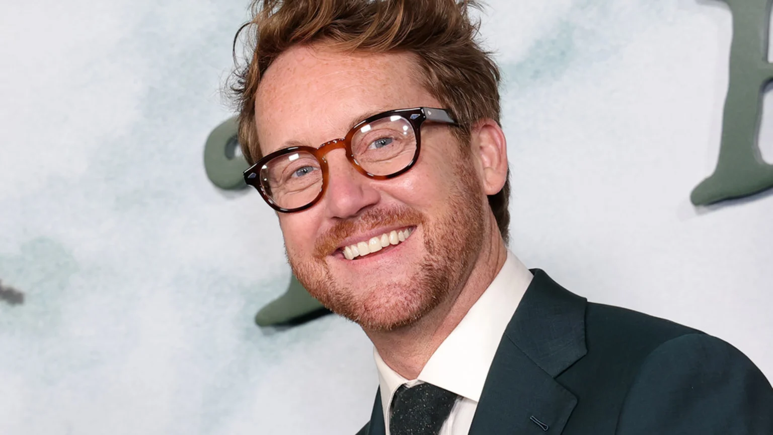A close-up portrait of a man in his 40s with reddish-brown hair and beard, wearing round tortoise-shell glasses, a dark suit jacket, white collared shirt, and dark patterned tie. He smiles broadly against a bright indoor event backdrop featuring large, blurry dark green shapes.