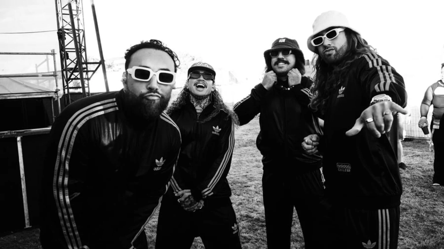 Four stylish men wearing matching Adidas tracksuits, sunglasses, and hats pose outdoors in a black and white photo.