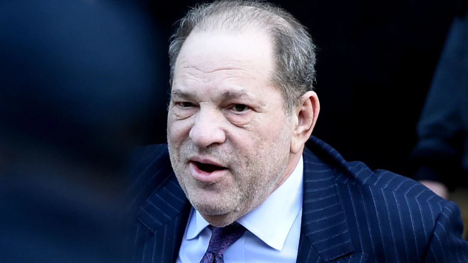 Close-up of Harvey Weinstein, an older man with light gray, thinning hair and a short beard, wearing a dark pinstripe suit and tie, with his mouth open as if speaking, against a blurred dark background.