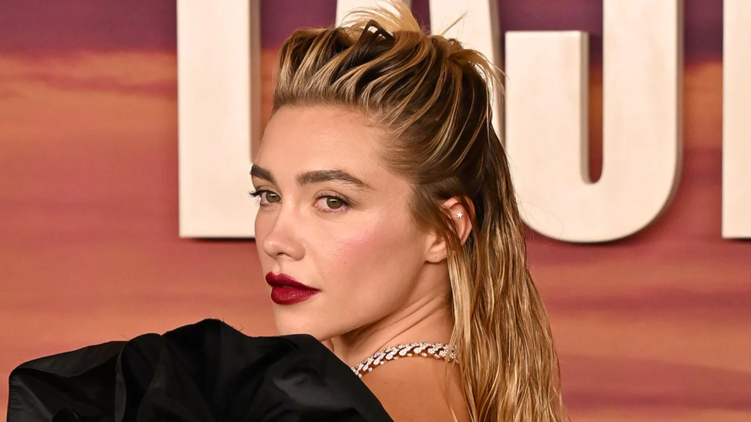 Florence Pugh looks over her left shoulder, wearing dark red lipstick, a star earring, a sparkly necklace, and a wet-look hairstyle, against a blurred, warm-toned background.