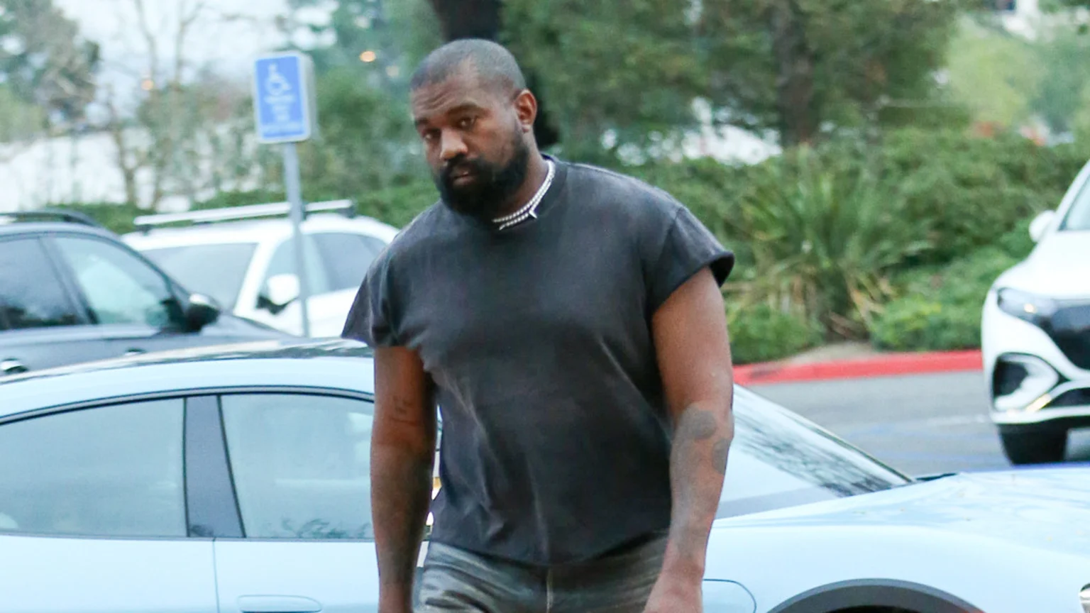 Kanye West, with a beard and short hair, wears a dark t-shirt and a beaded necklace while looking towards the left, with cars and greenery in the background.