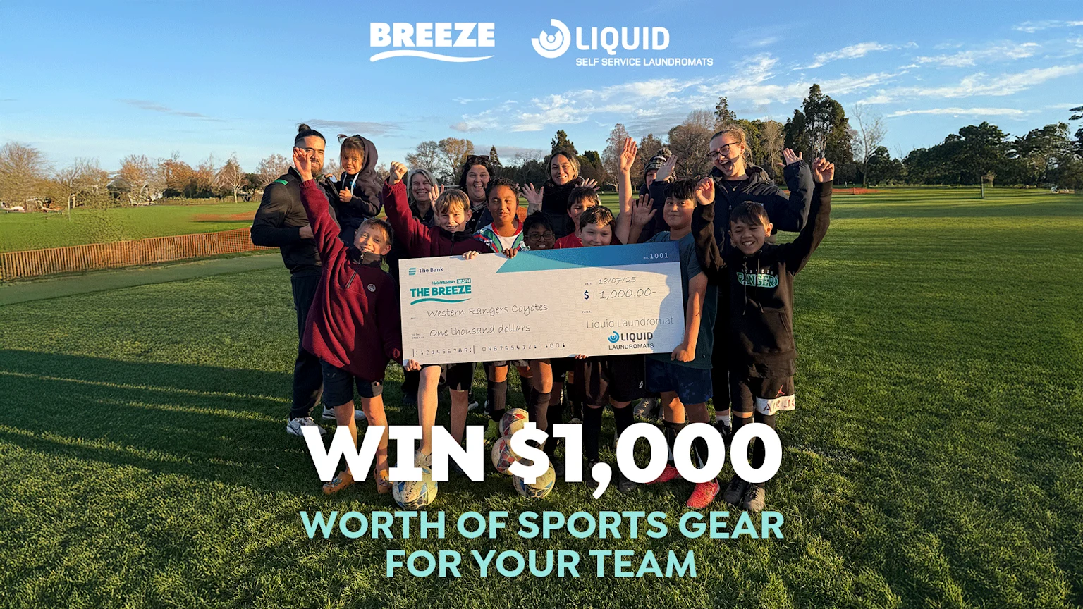 A group of happy children and adults celebrate on a grassy field, holding a large check for $1,000 from Breeze and Liquid Laundromats for sports gear, with company logos visible at the top.