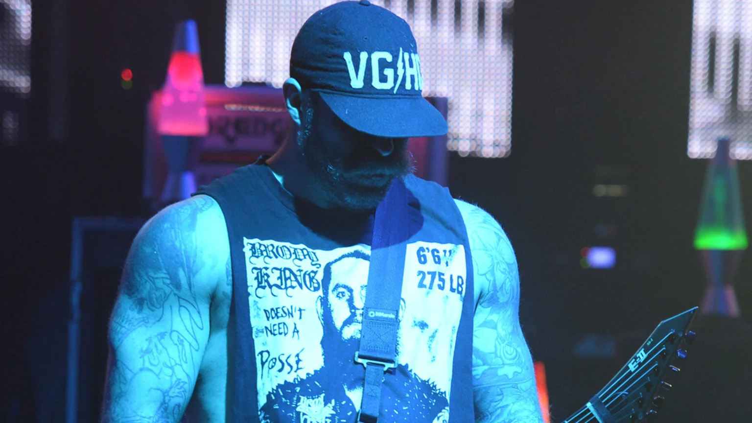 A bearded man with tattoos wears a 'VG/H' cap and a sleeveless shirt with 'BROTHAL KING' text, a bearded face, and '6'6" 275 LB', holding a guitar. He is under blue light on a dark stage with lava lamps.