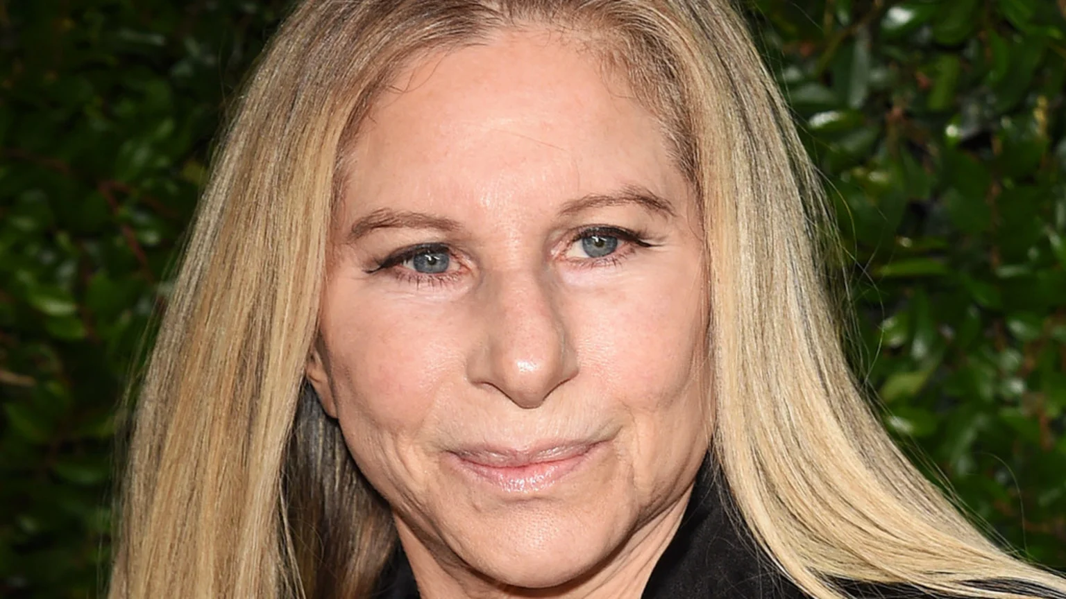 A close-up portrait of Barbra Streisand, a woman with long blonde hair, blue eyes, and fair skin, looking towards the camera with a slight smile against a blurred green background.