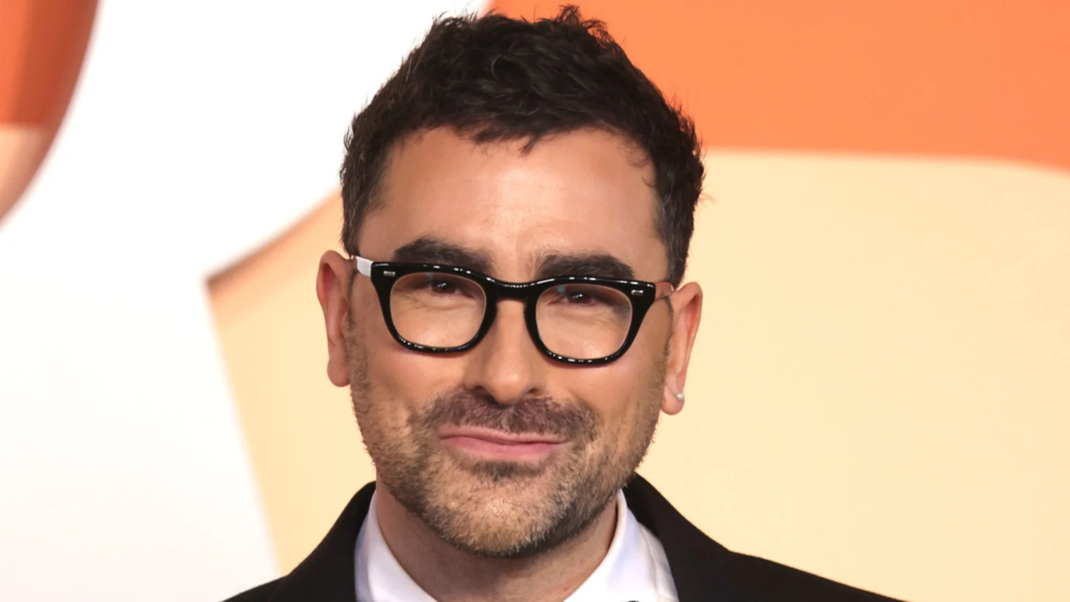 A man with dark wavy hair, a beard, and black glasses with white accents smiles slightly at the camera, wearing a dark suit jacket against a warm-toned background.