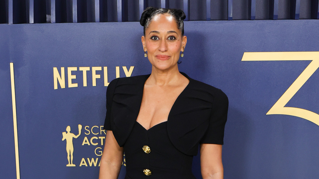 Tracee Ellis Ross poses, looking towards the viewer, wearing a black V-neck dress with gold buttons and her hair in two buns, against a blue wall with gold "Netflix" and "SCR ACT G AW" text.