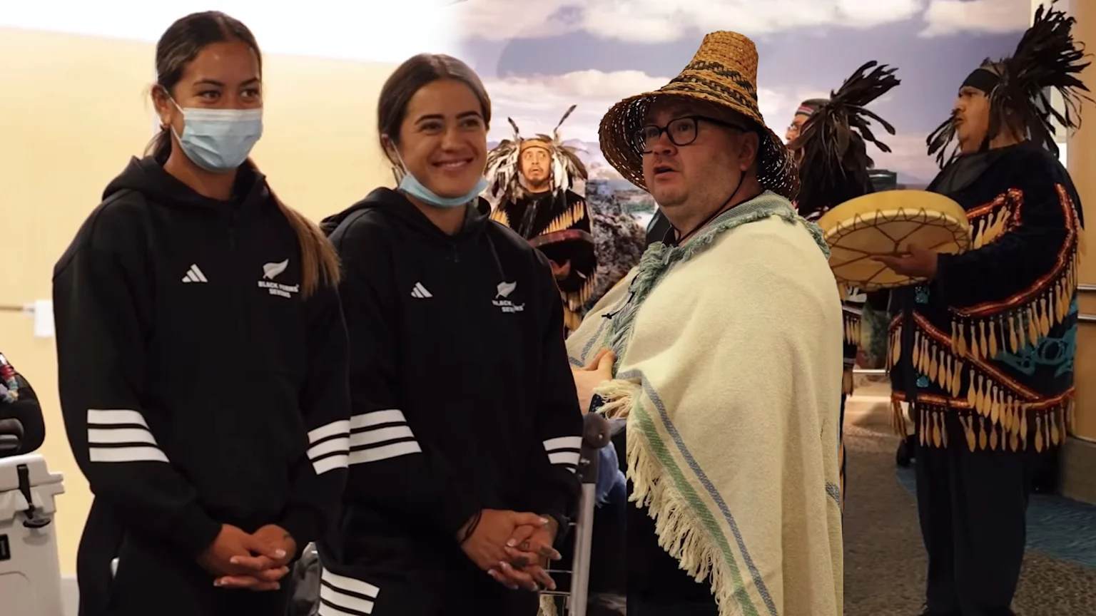 Two women in black Black Ferns Sevens hoodies stand left, observing a man in a straw hat and shawl, with Indigenous individuals in traditional attire and a drummer in the background.