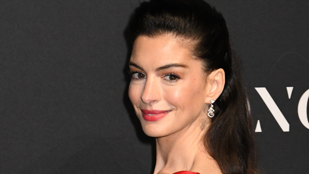 Close-up of Anne Hathaway smiling and looking at the camera, with her hair pulled back, wearing earrings and red lipstick against a dark background with white lettering.
