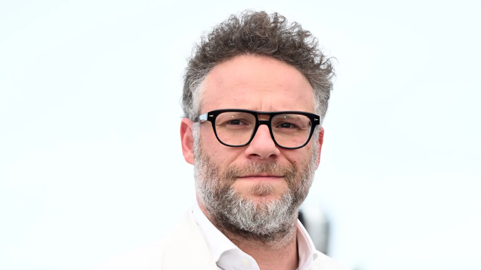 A man identified as Seth Rogen with curly graying hair, a full gray beard, and black-framed glasses smiles faintly while looking directly at the camera, wearing a white shirt and light-colored jacket against a bright, out-of-focus background.
