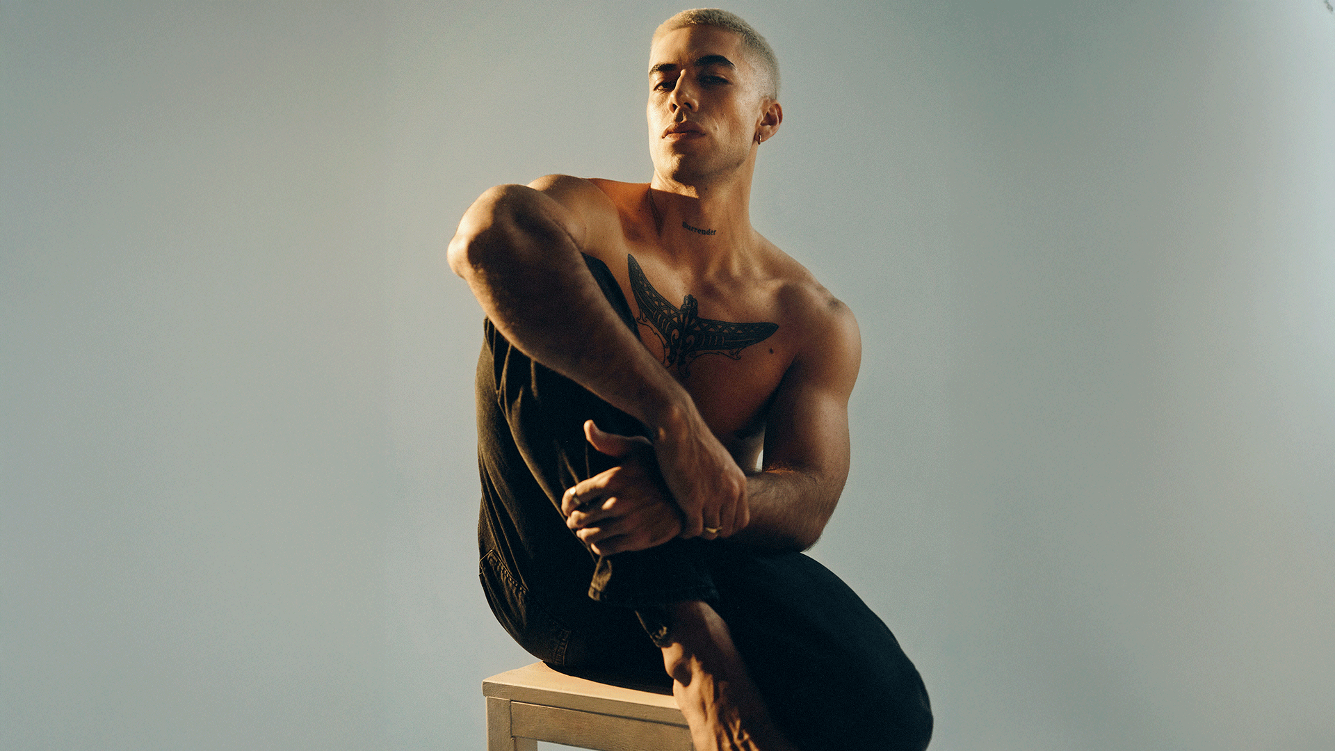 A shirtless man with short light hair and chest tattoos sits on a light stool, looking at the viewer, with his hands resting on his knee.