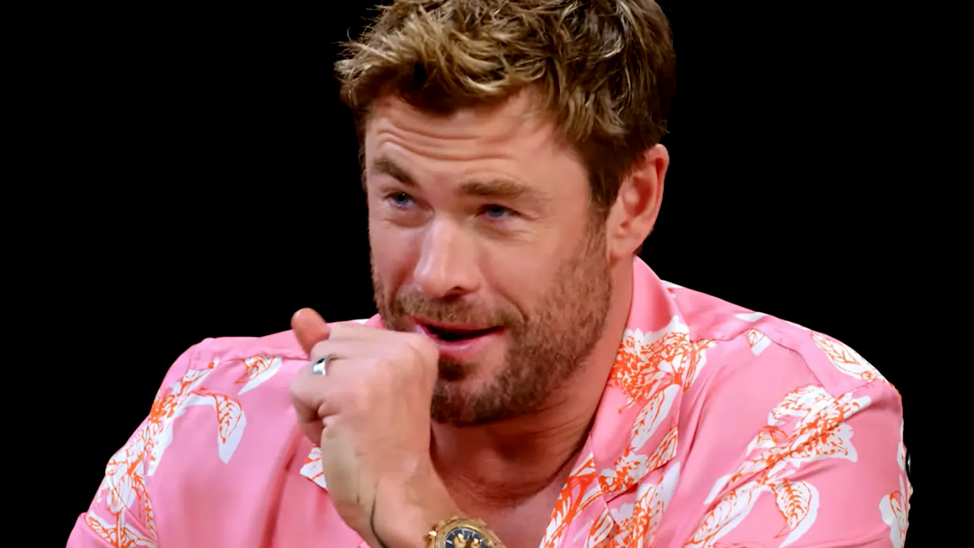 Close-up of Chris Hemsworth looking directly at the camera with blue eyes, short brown hair, and a light beard, against a blurred light grey background with faint text.