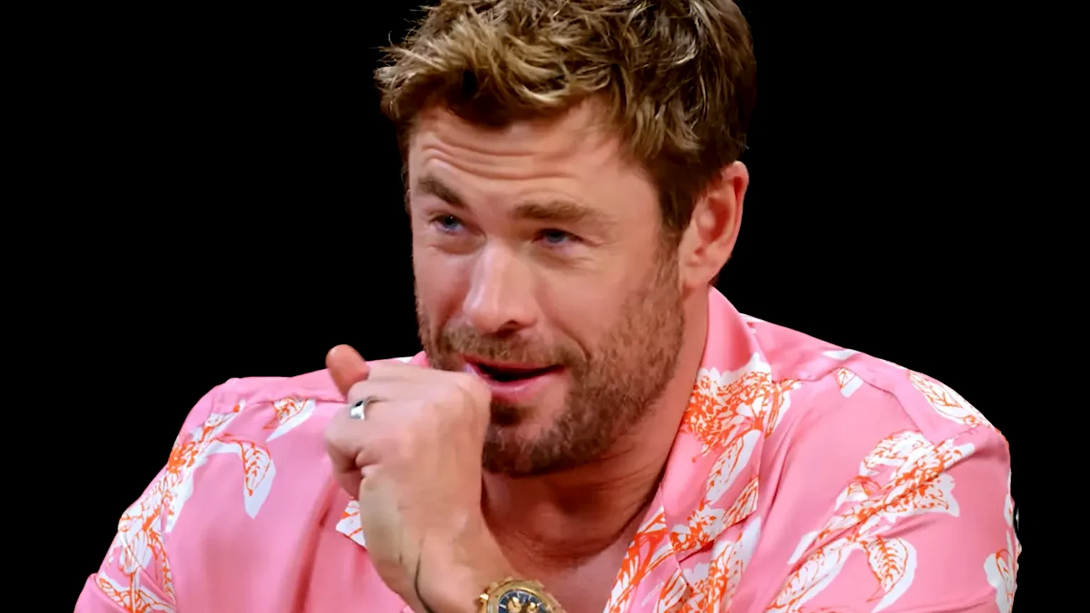 Close-up of Chris Hemsworth looking directly at the camera with blue eyes, short brown hair, and a light beard, against a blurred light grey background with faint text.