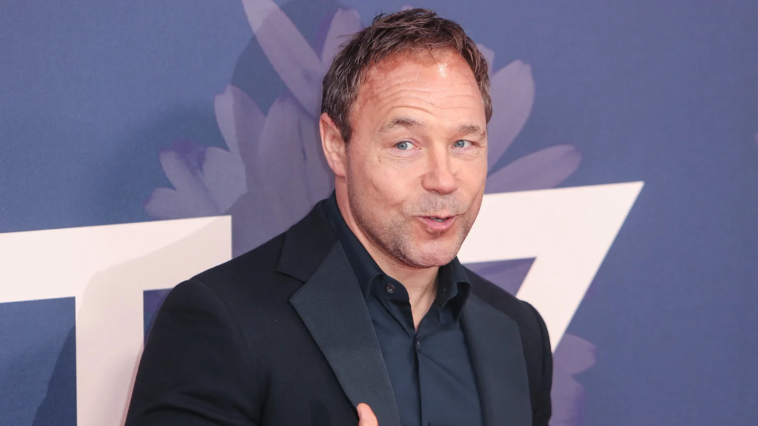 A man with short dark hair and blue eyes, wearing a dark suit jacket and shirt, stands against a blue backdrop with white geometric and faint floral patterns, his mouth slightly open.