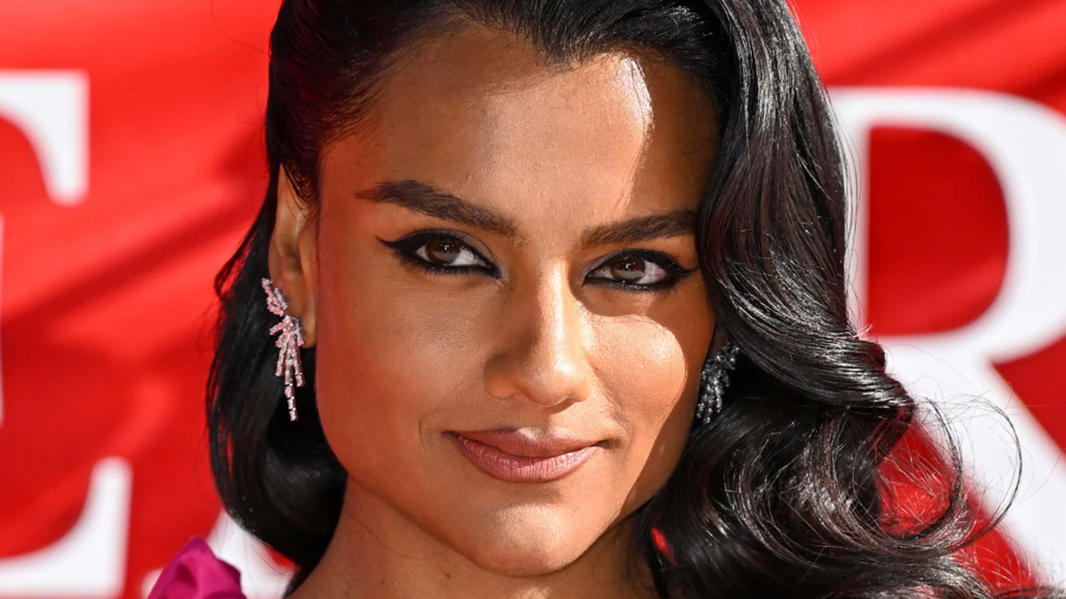 A smiling woman with dark skin, dark wavy hair, winged eyeliner, and sparkling earrings poses against a blurry red background with white text.