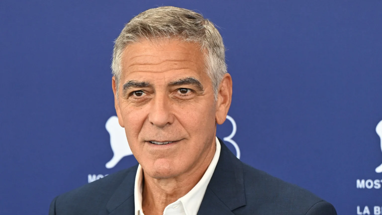 George Clooney, with grey hair and a slight smile, is seen from the chest up against a blue background, wearing a dark suit jacket and white shirt.