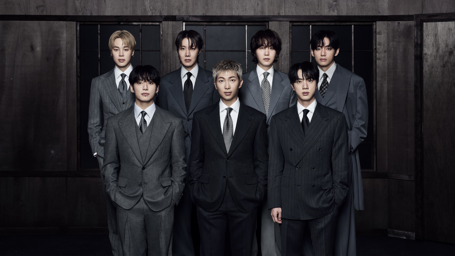 Seven members of BTS stand staggered in various dark suits against a dark wooden wall.