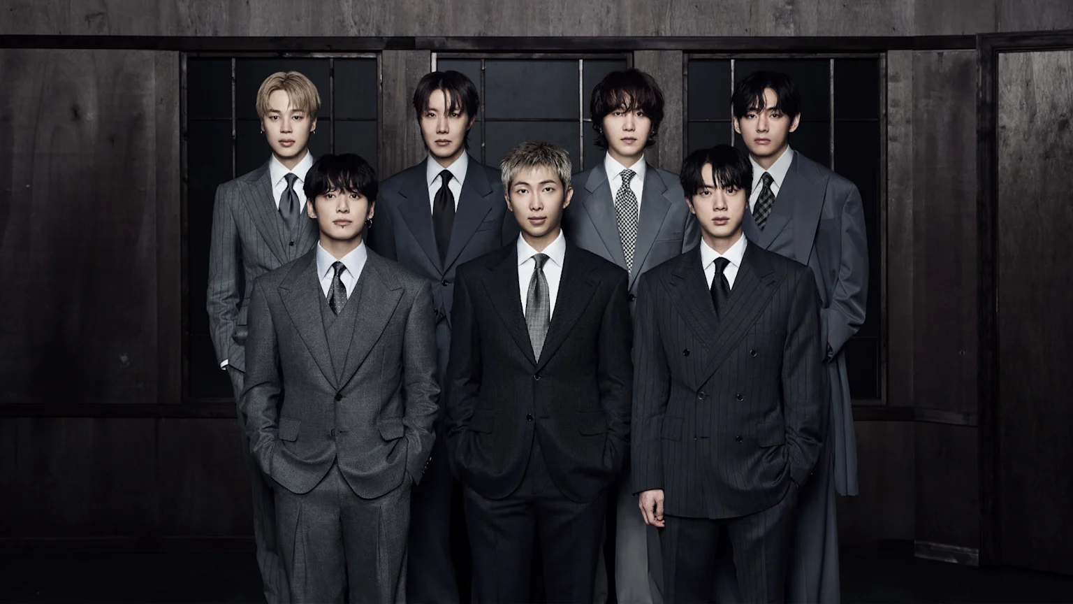 Seven members of BTS stand staggered in various dark suits against a dark wooden wall.