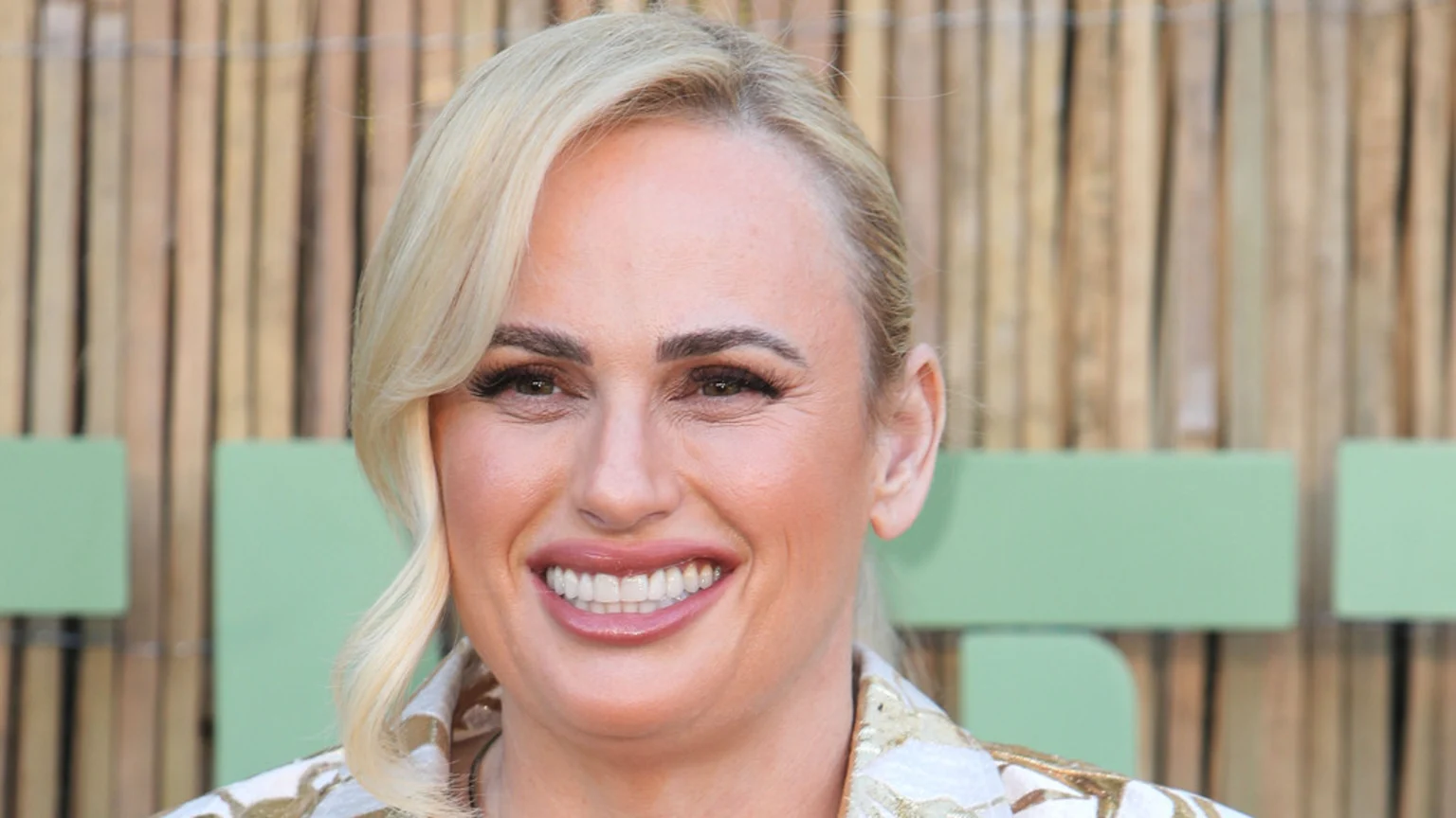 Close-up of Rebel Wilson smiling directly at the camera, with blonde hair, visible makeup, and a white and gold patterned jacket, against a blurred background.