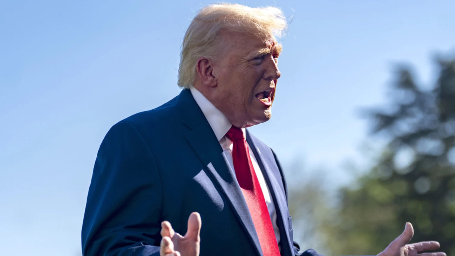 Donald Trump, wearing a blue suit and red tie, speaks outdoors with his mouth open and hands gesturing, against a clear blue sky and blurred trees.