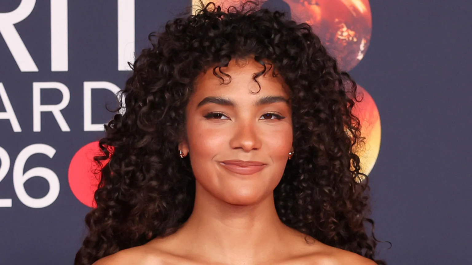 A smiling woman with long, dark curly hair looks at the camera, against a blurred dark background with white text "BRIT AWARDS 26" and red/orange circular elements.