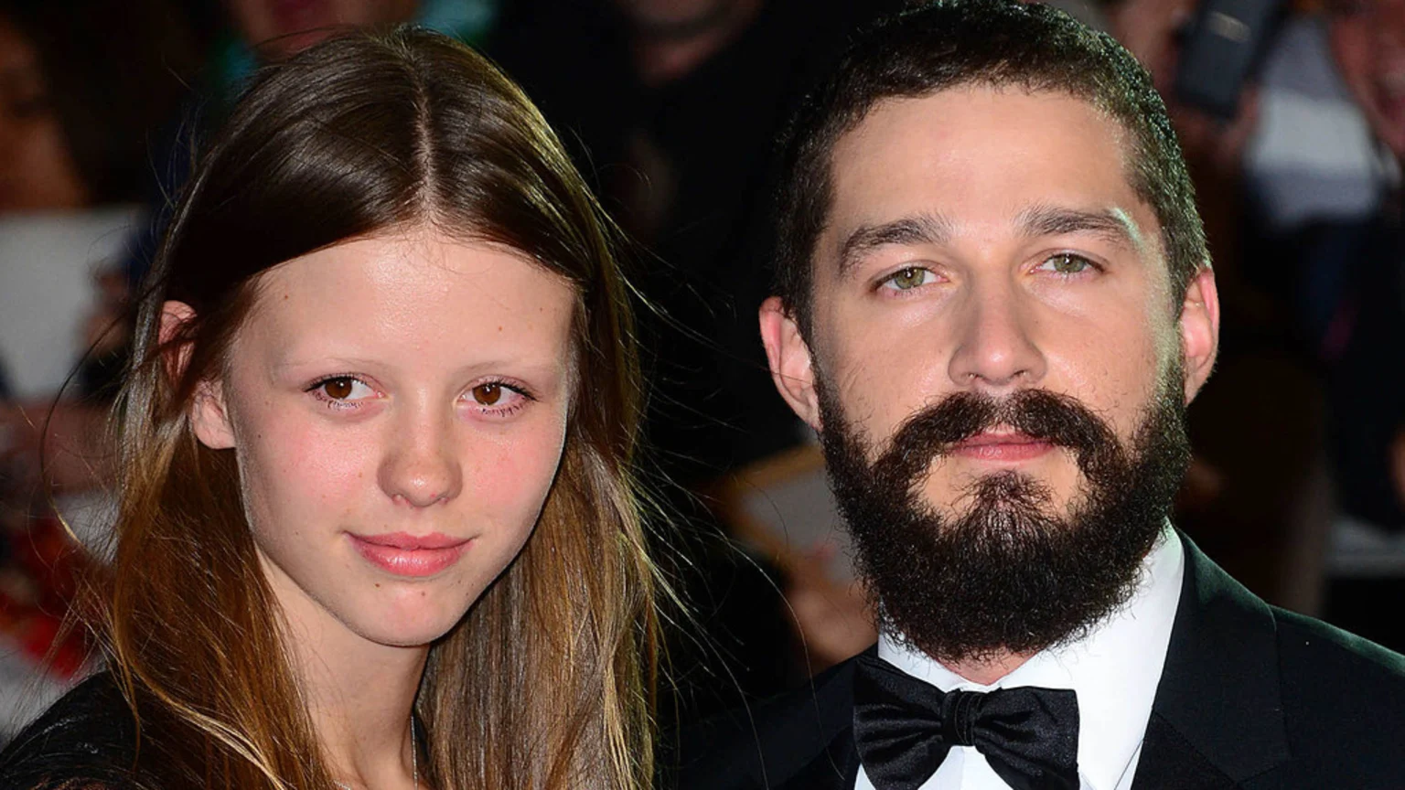 A close-up of actress Mia Goth with long brown hair and actor Shia LaBeouf with a beard, mustache, and bow tie, both looking forward.