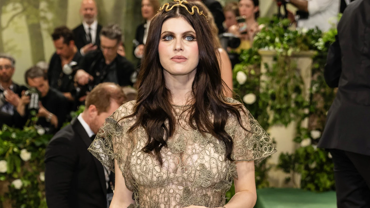 Alexandra Daddario, with long dark hair and striking blue eyes, wears a golden snake-like headband and a sheer gold lace top, posing in front of a blurred crowd and green foliage.