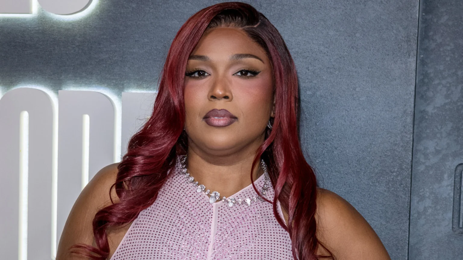 Lizzo, a Black woman with long reddish-brown hair and a pink sparkling top, looks directly at the camera, wearing a diamond necklace against a dark gray background with partial white letters.