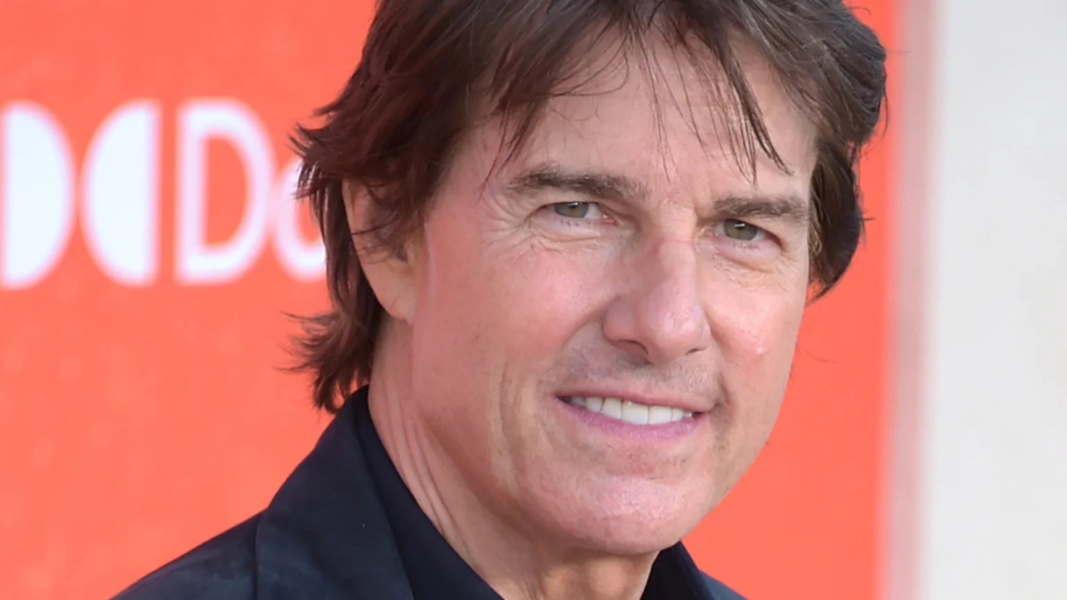 Close-up of actor Tom Cruise smiling, wearing a dark shirt, against a blurred orange background with a partially visible white "DOLBY" logo.