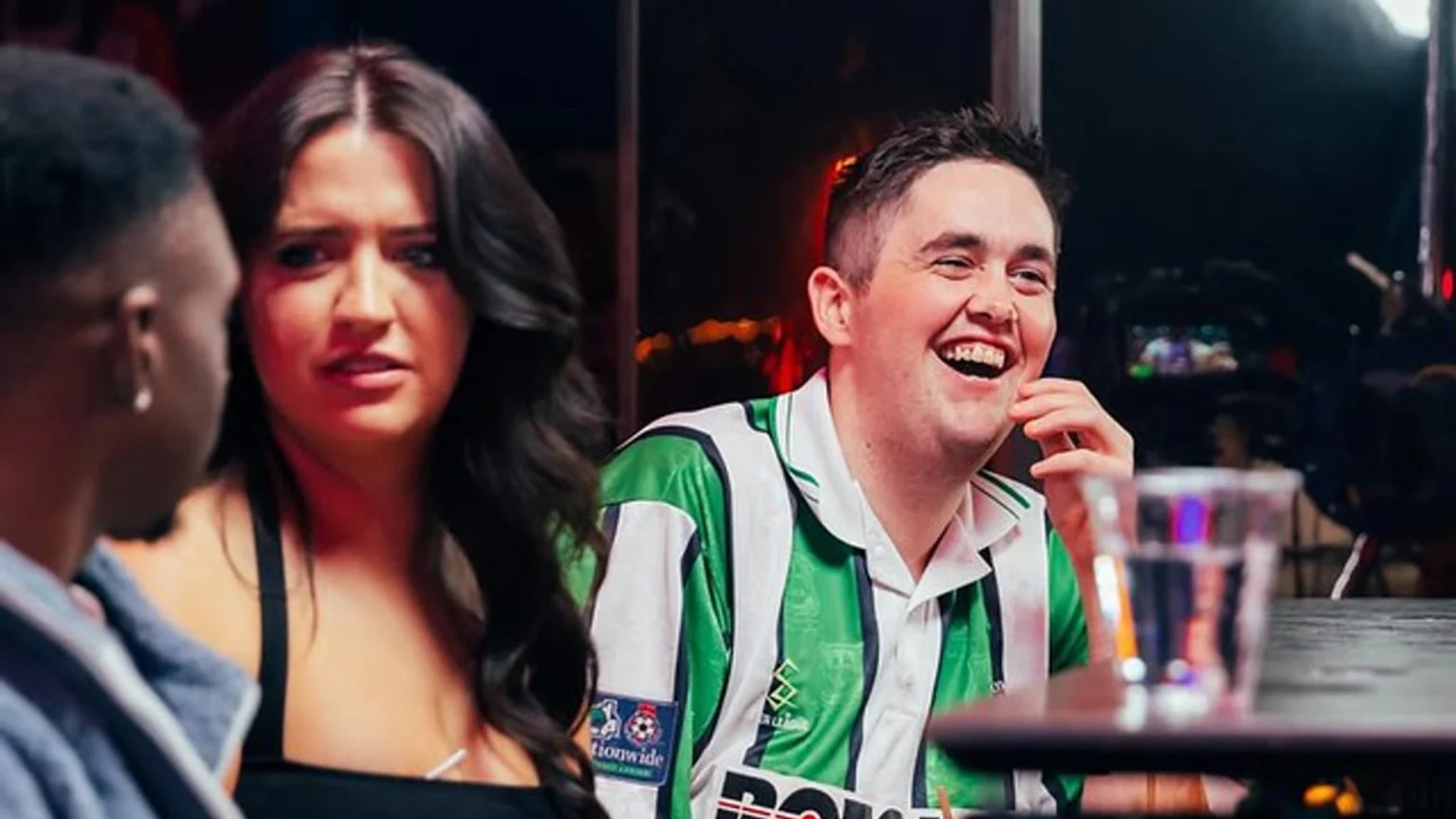 A man in a green and white striped shirt laughs heartily while a woman next to him looks concerned, with a third person partially visible on the left in an indoor, possibly filmed, setting.