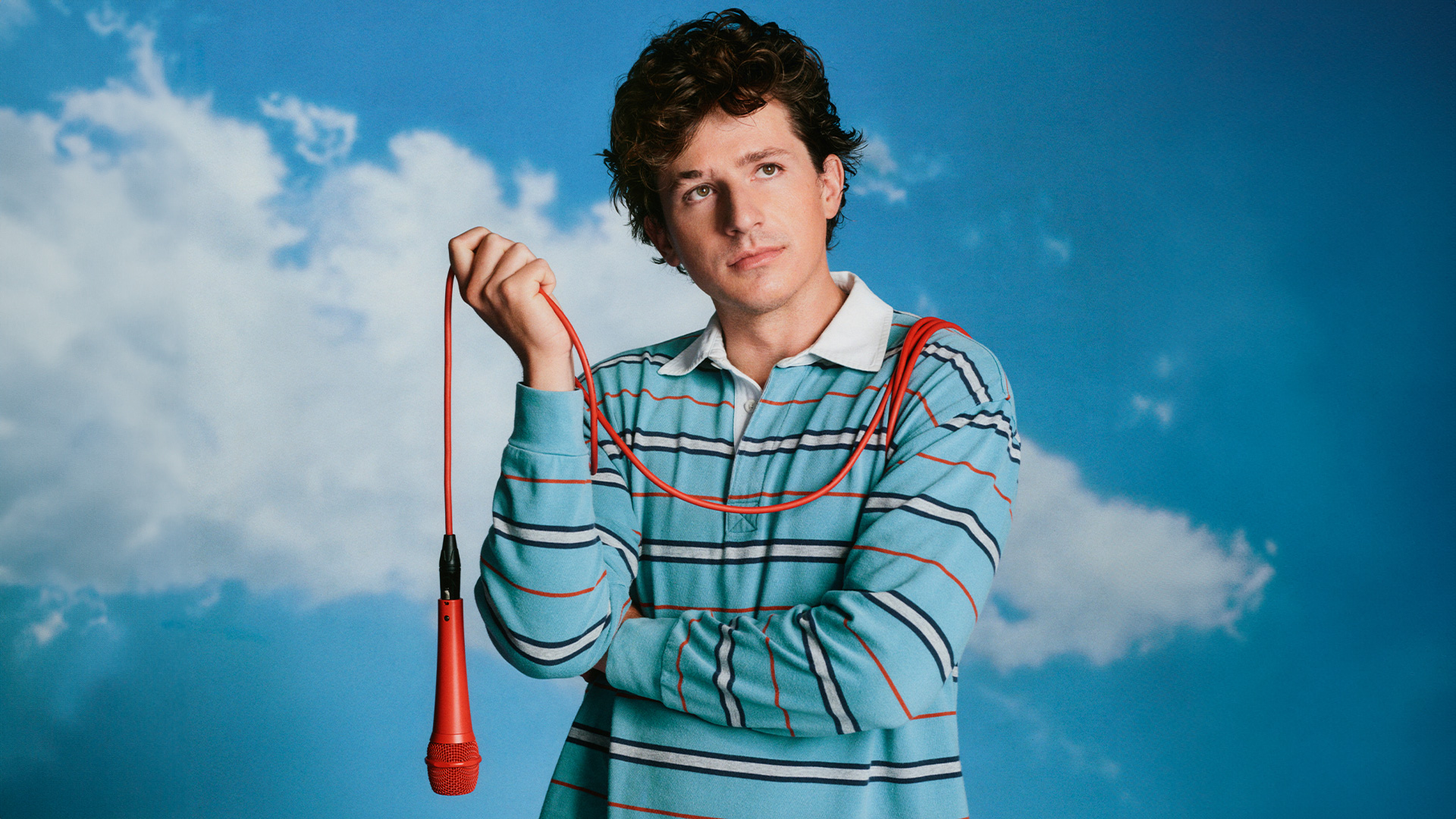 Charlie Puth holding a red-orange microphone against a sky backdrop