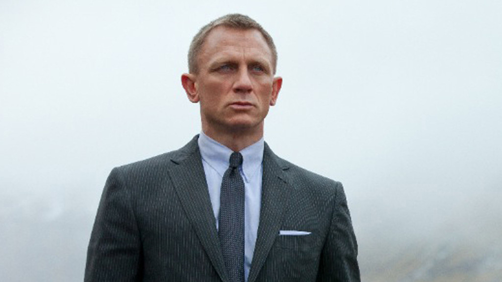 A man with short hair and a serious expression wears a dark pinstripe suit, light blue shirt, dark tie, and white pocket square against an overcast sky.