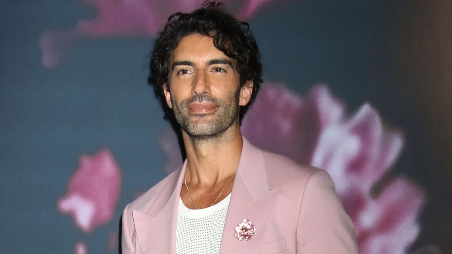 A man with dark curly hair and a beard, wearing a pink blazer with a floral brooch over a white shirt, stands against a blurred blue background with pink floral shapes.